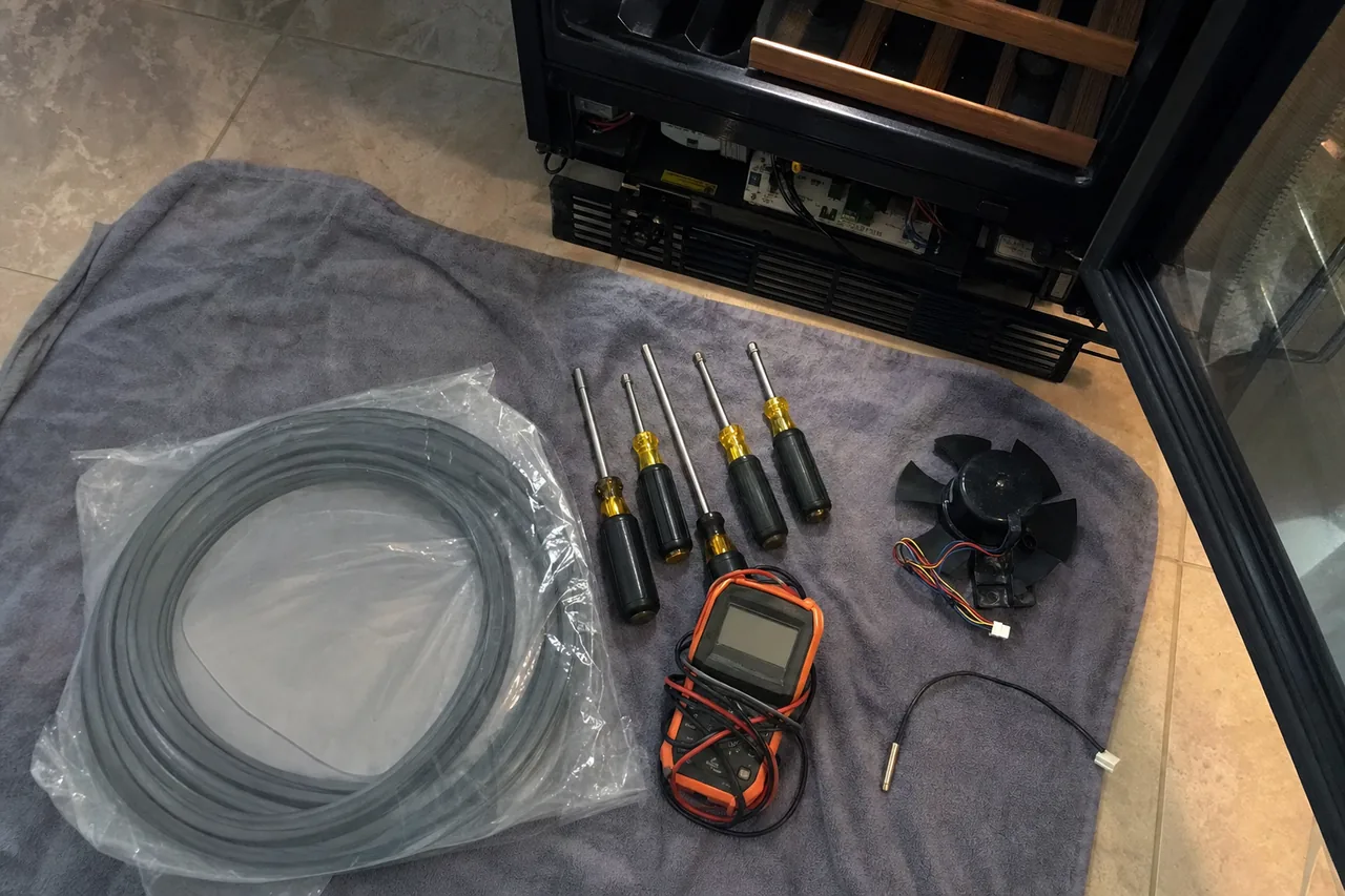 An overhead flat-lay on a grey towel of nut drivers, a multimeter, a coiled replacement door gasket in a bag, a fan motor and a thermistor probe, laid beside the open base of a wine cabinet.