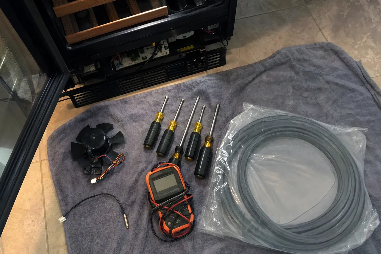 The open lower base of a wine cabinet with its control board and fan bay exposed, tools laid on a towel on the tiled floor in front of it.