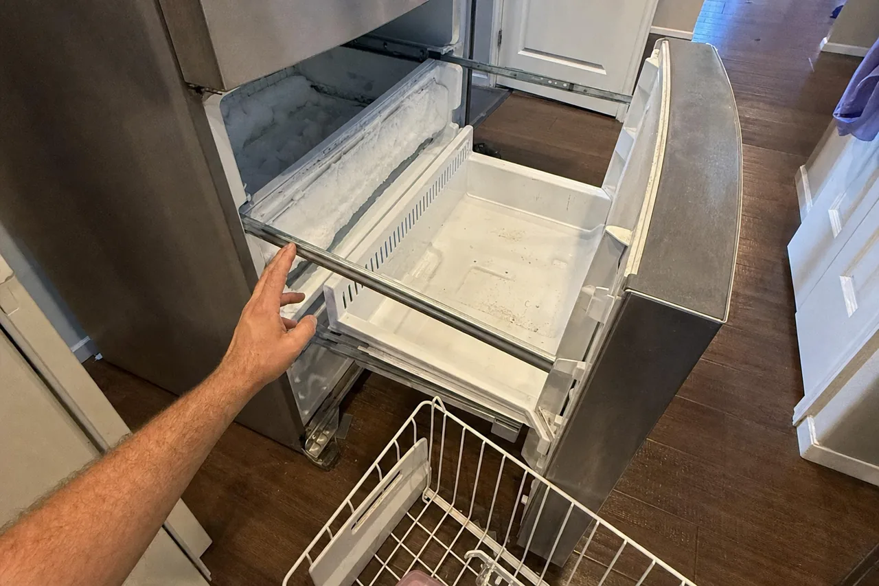 A bottom freezer drawer pulled fully open on its runners, frost built up along the rear wall and the wire basket lifted out onto the floor.