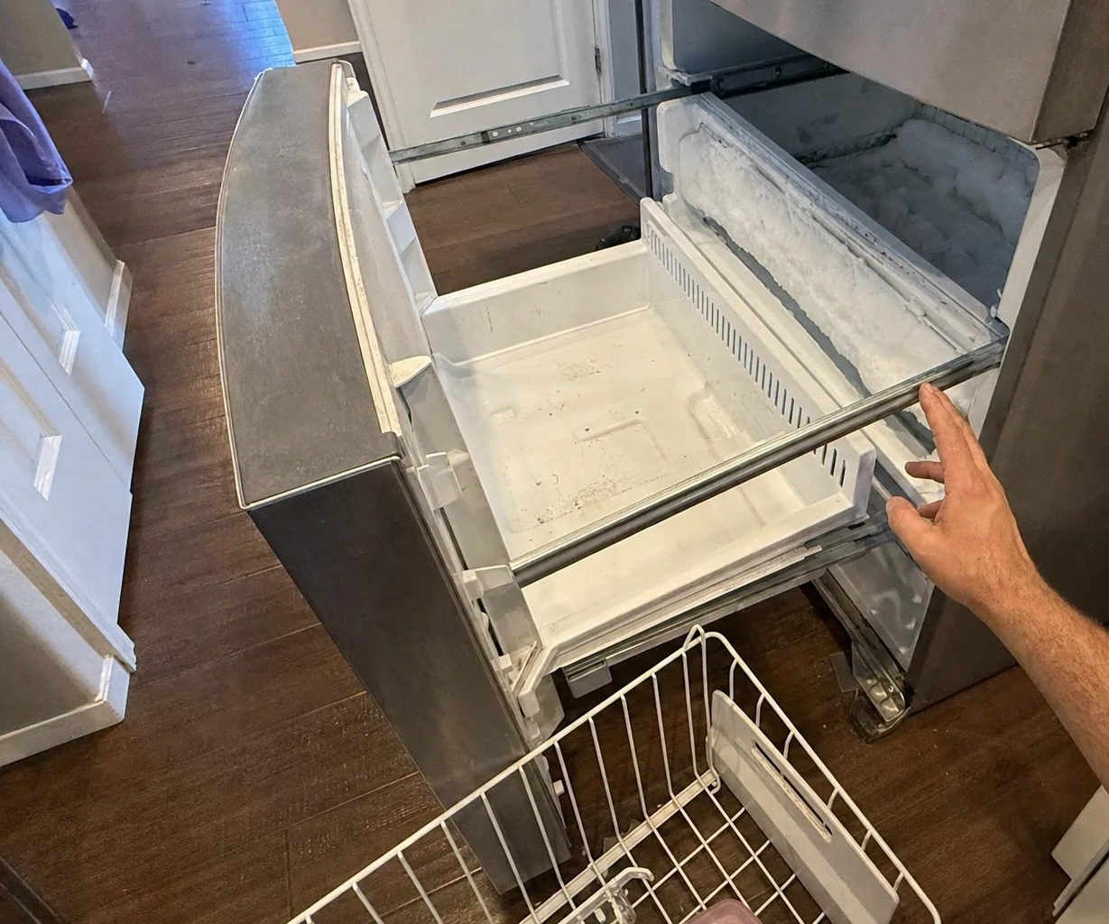 Hands checking the runner track of an open freezer drawer, frost visible along the gasket above it.