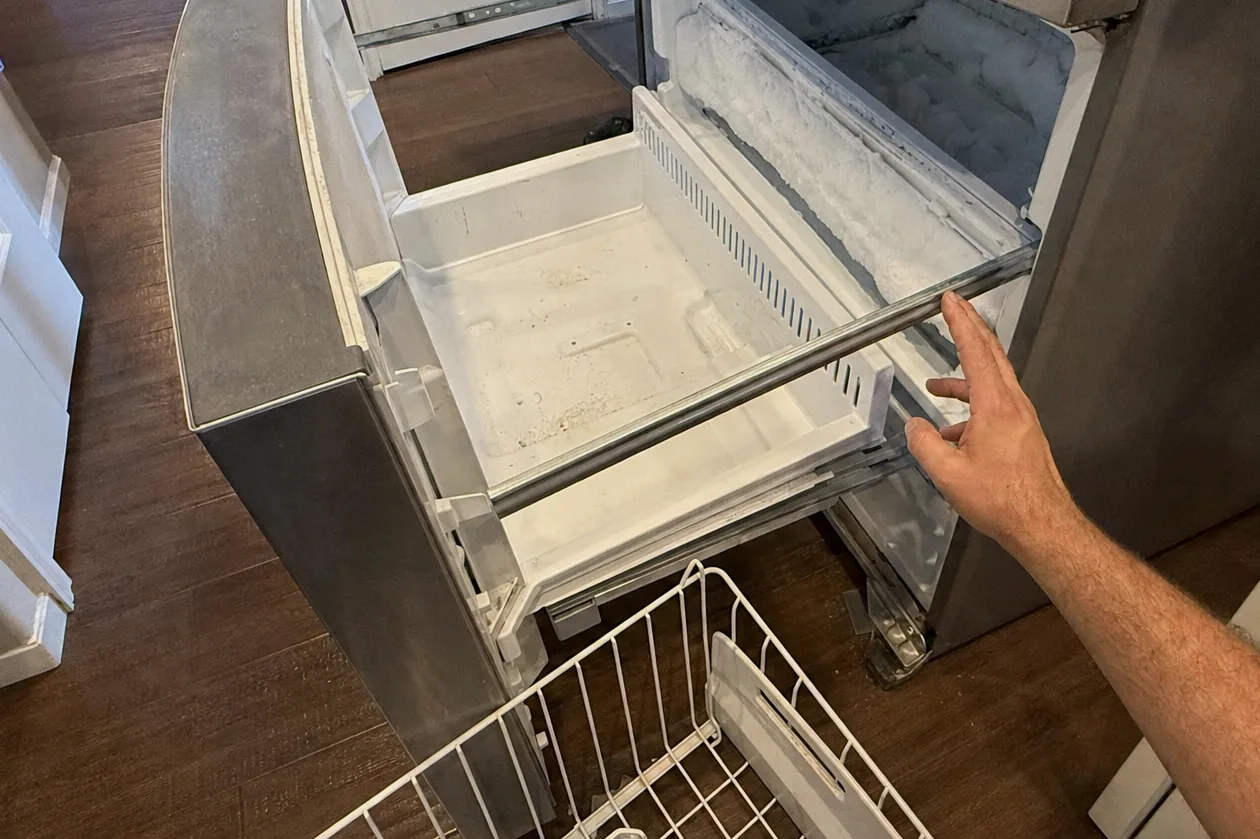 A freezer drawer standing open on a kitchen floor, wire basket set down beside it mid-job.