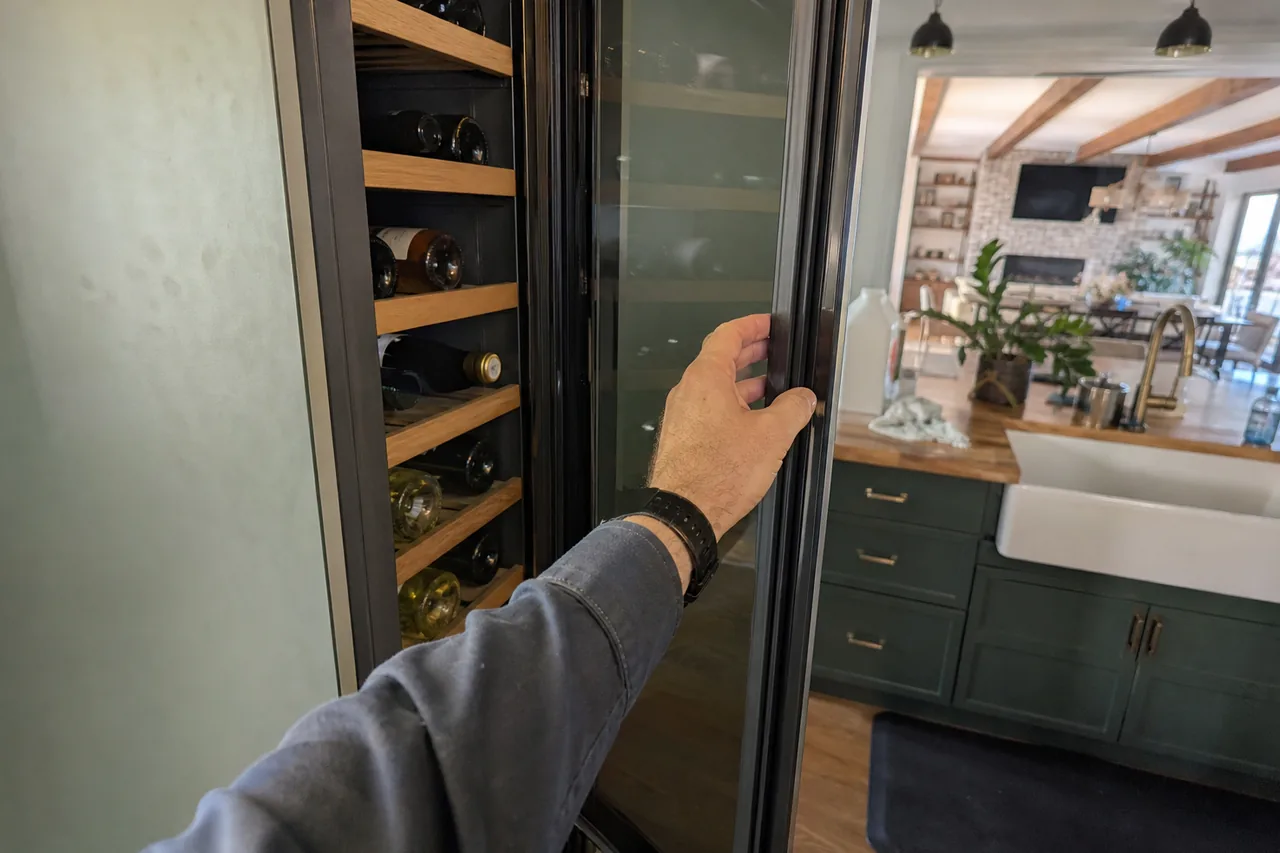 A hand in a grey sleeve pulls open a glass wine column door beside wooden racks of bottles, with green cabinets and a farmhouse sink in the kitchen behind.